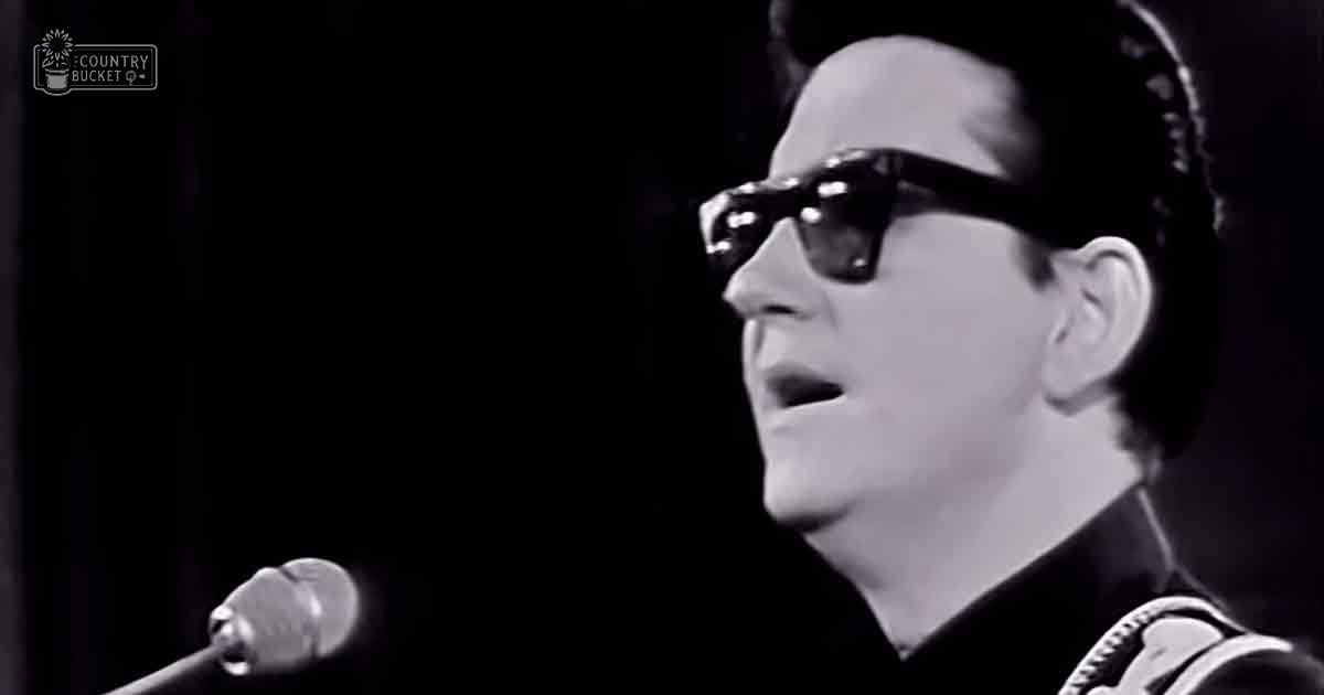 Adult male singer wearing dark sunglasses and a high-collared garment with rope-like detail on shoulder strap, singing into a microphone in a dimly lit studio setting.