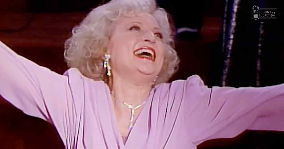 Older adult female in loose-fitting light purple blouse with deep V-neckline and large dangling earrings performing under stage lighting.