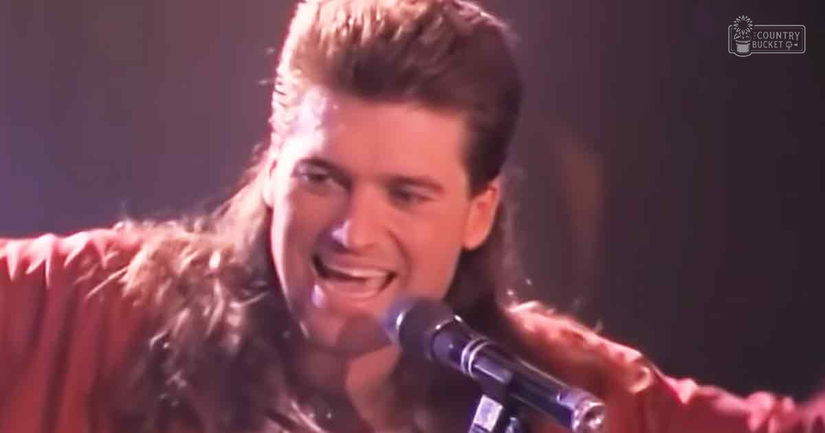 A young adult male singer wearing a red-brown, textured fur-lined jacket with a high collar and long sleeves performs on stage under bright spotlights against a dark background.