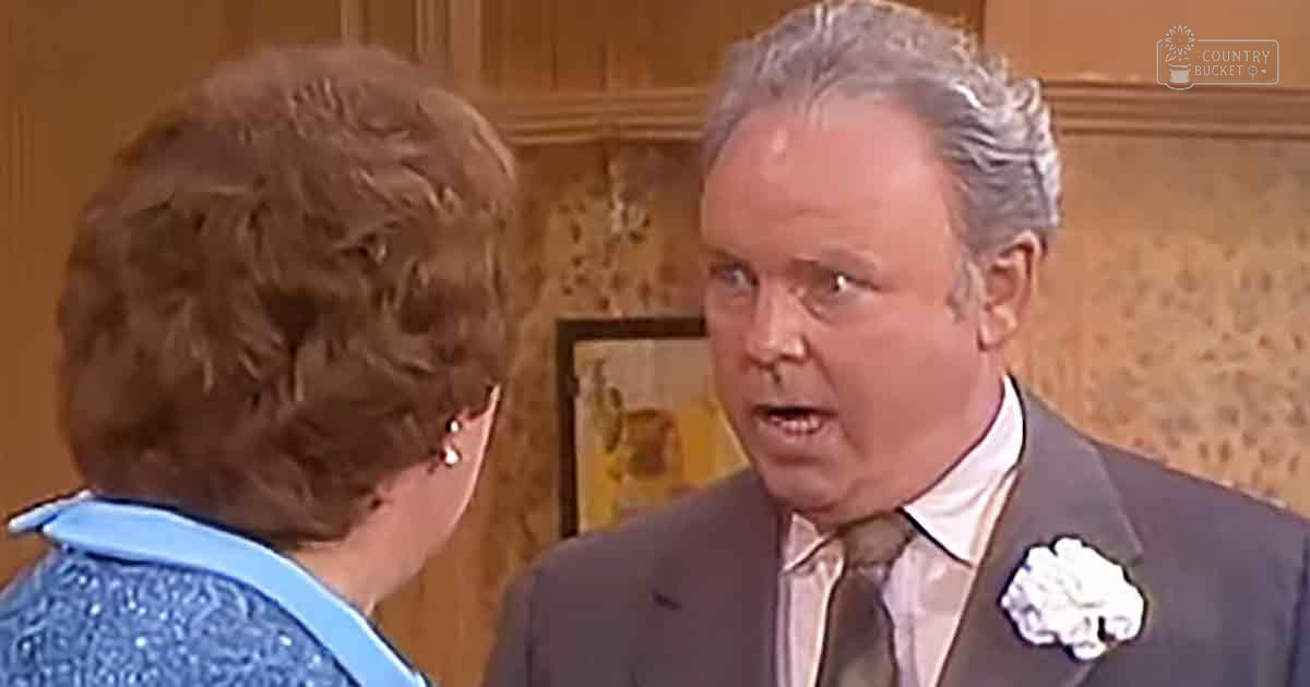 A middle-aged man wearing a dark gray suit jacket with a white dress shirt and light-colored tie, conversing with an older adult woman dressed in a blue patterned blouse and glasses, set against wooden paneling and framed artwork. The scene suggests a formal or festive occasion.