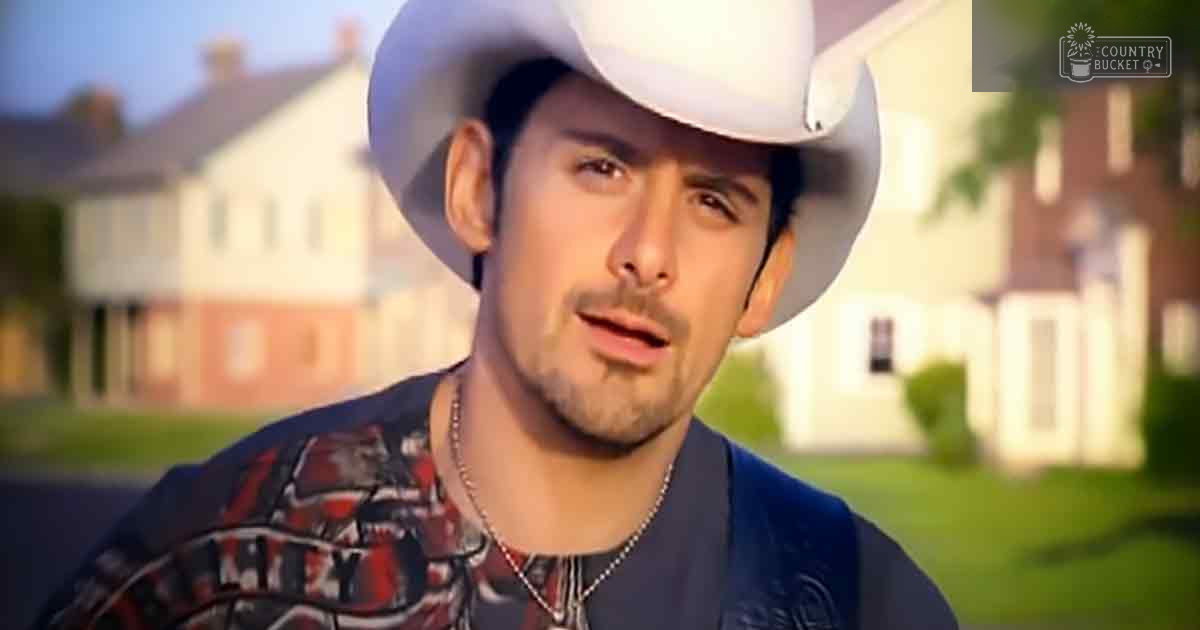 A young adult male in a suburban neighborhood, wearing a red and black patterned cowboy shirt with a blue jacket over it, white cowboy hat, surrounded by houses with brick facades and greenery. The setting suggests spring or summer due to the lush vegetation.