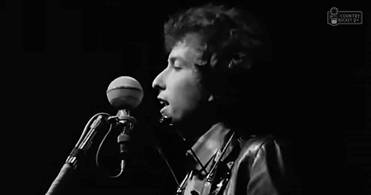 A young adult male singer wearing a dark leather jacket performs on stage with a microphone and guitar strap visible.