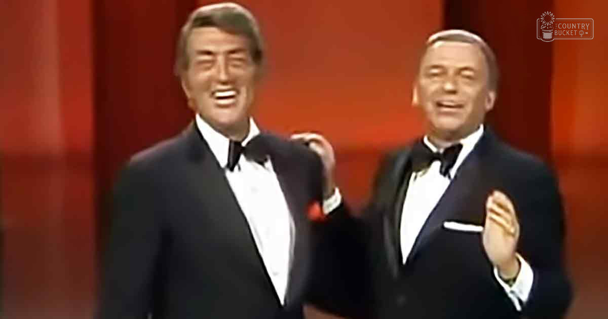 Two adult males dressed in classic tuxedos perform on a stage with red curtains, suggesting an indoor venue for a formal event.