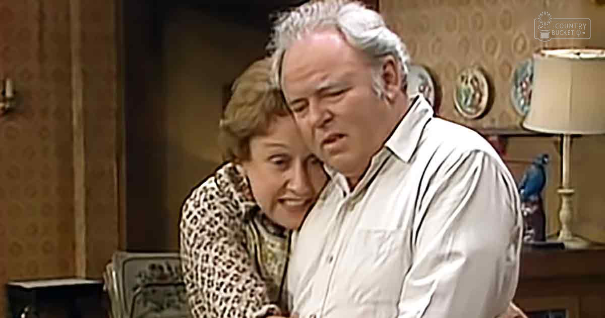 A middle-aged man in a white button-up shirt with vertical stripes stands beside a middle-aged woman wearing a leopard print top in an indoor setting. The decor suggests an older style living room, possibly from the mid-20th century.