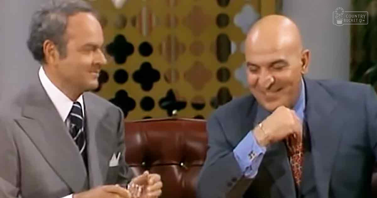 Two middle-aged to older adult men in a studio setting, wearing formal suits and accessories, seated with a patterned wall background suggesting an indoor studio environment.