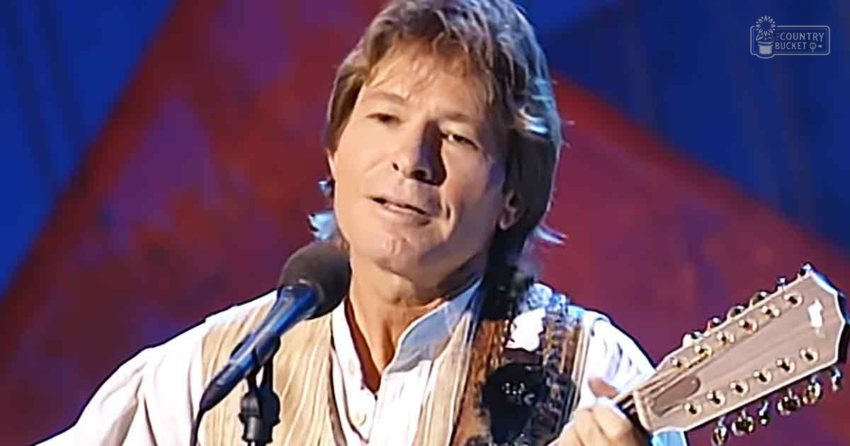 A middle-aged adult male wearing a relaxed fit, white button-up shirt with vertical stripes and a collar, holding an acoustic guitar while performing at an indoor event under red stage lighting.