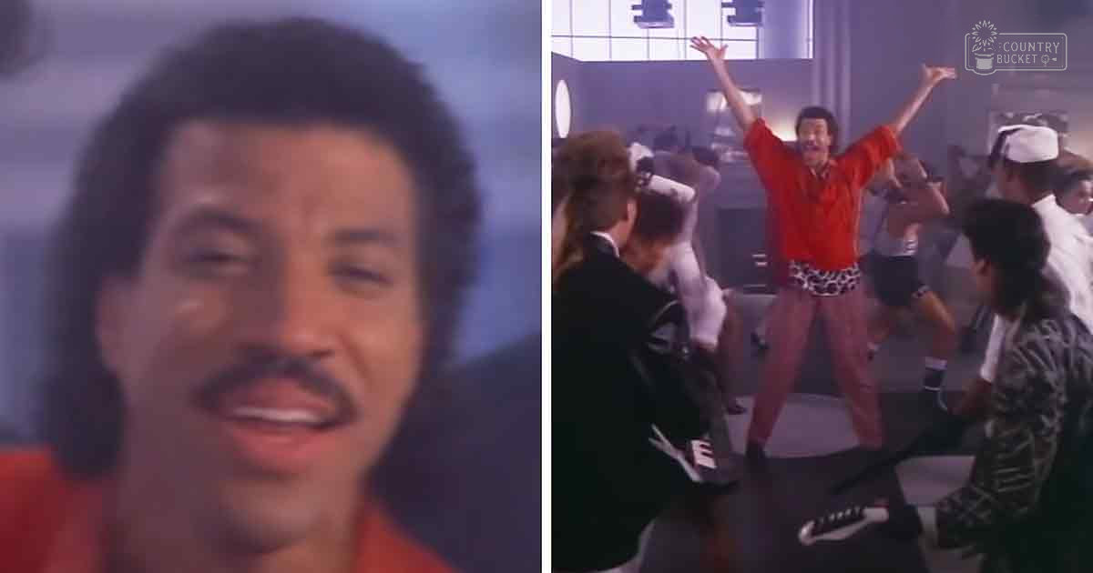 An adult male singer with long hair, wearing an orange shirt and leopard-print pants, smiles widely while performing in a dance studio filled with mirrors and natural light.