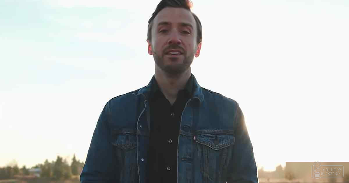 A young adult male in a dark blue denim jacket and black shirt stands outdoors with trees and a clear sky, suggesting early morning or late afternoon sunlight.
