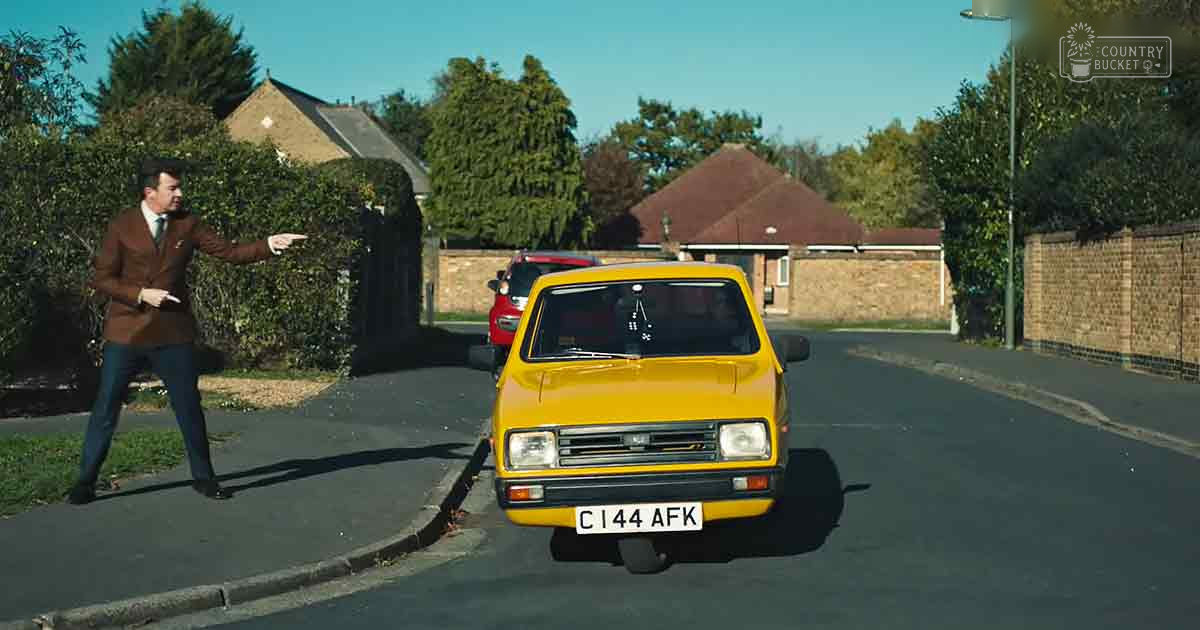 Adult male in urban residential area, wearing brown patterned blazer with dark trousers, black shoes; yellow car prominent in foreground.