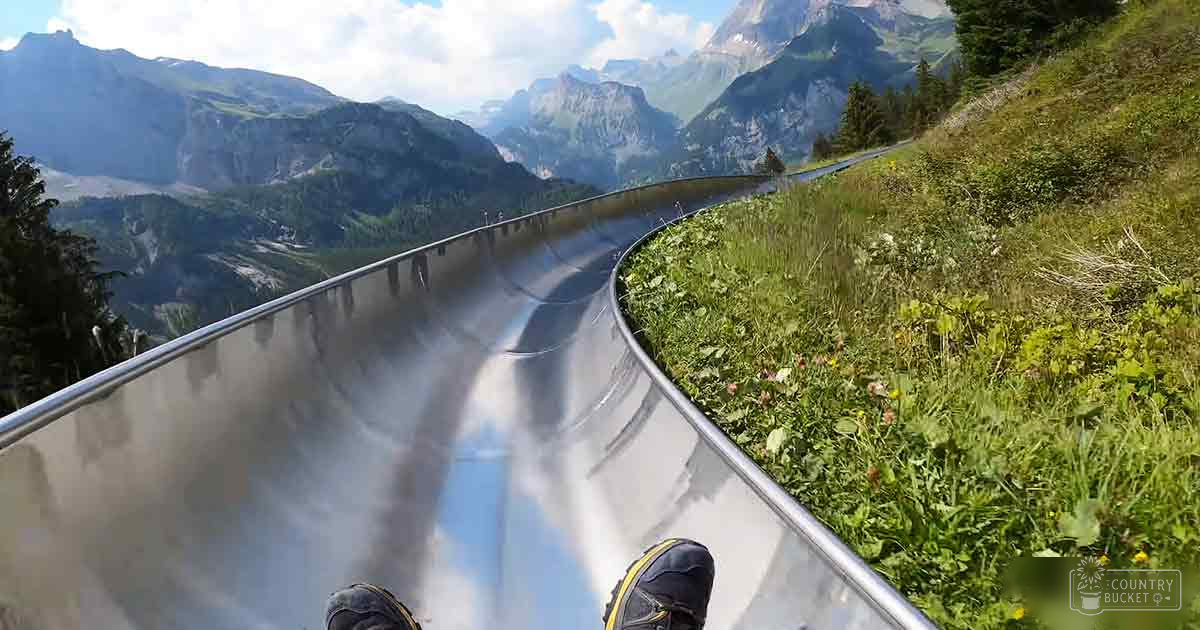 Feet wearing black shoes with yellow accents sliding down an alpine slide in Switzerland on a partly cloudy day during late spring or summer.