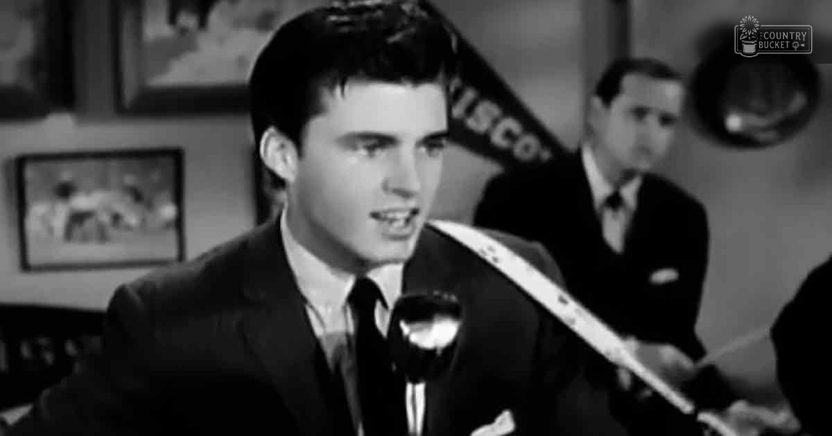 Young adult male wearing a dark suit jacket, white dress shirt with black tie and pocket square in an indoor studio setting.