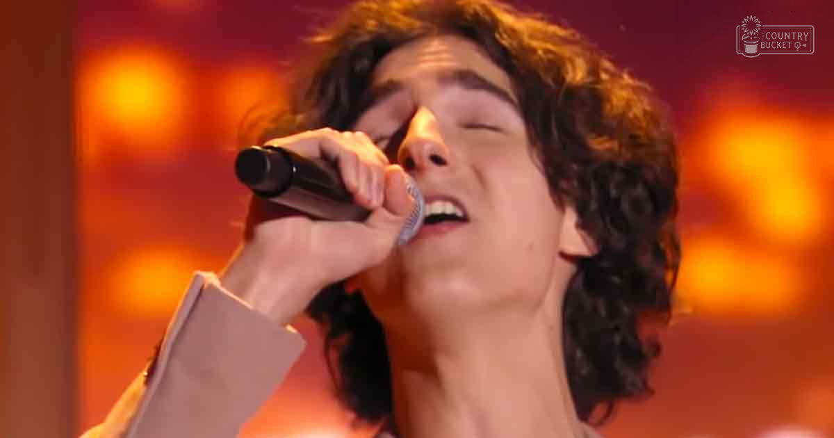 A young adult male singing into a microphone on stage under bright orange and red lighting.