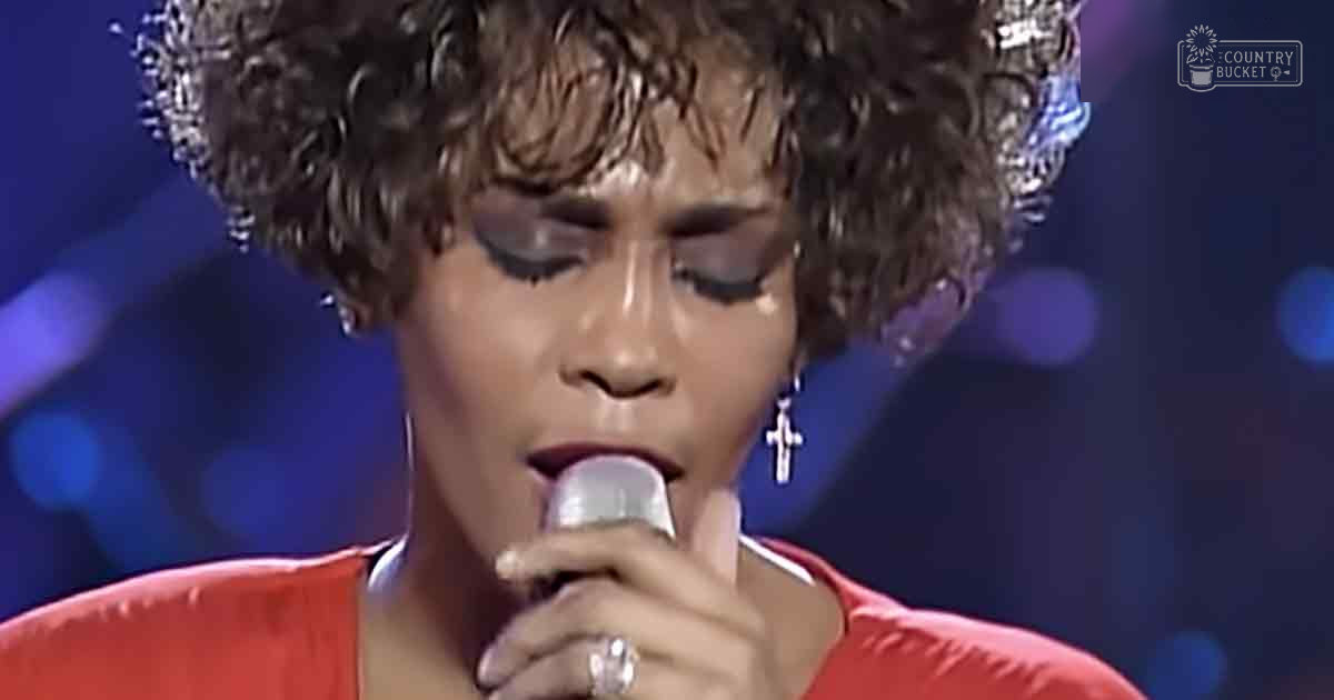 A female adult singer in a red, silk-like 1980s or early 1990s garment with short sleeves and a round neckline, wearing a ring on her left hand, earrings with a cross design, and a tiara-like headpiece, singing into a microphone against dark blue stage lighting.