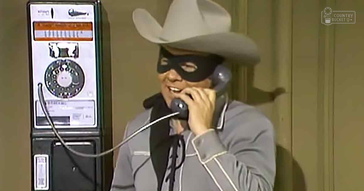 A man in a Western-style costume, including a cowboy hat and mask, interacts with a vintage payphone indoors.