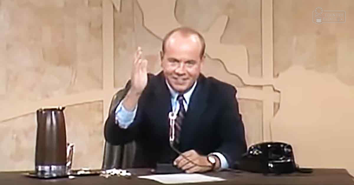 Middle-aged to older adult male wearing a dark suit jacket, light blue shirt, and patterned tie, standing indoors in what appears to be a news studio or office setting.
