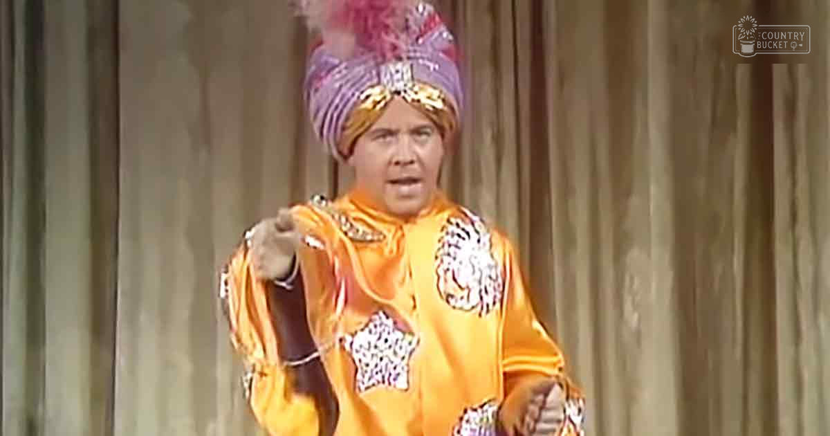 An adult male performer in a vibrant orange robe with intricate patterns and a turban adorned with feathers, beads, and other decorative elements on stage.