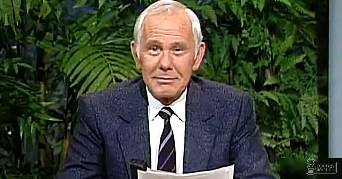 Older adult male wearing a formal suit and tie, standing in a studio setting with green foliage in the background.