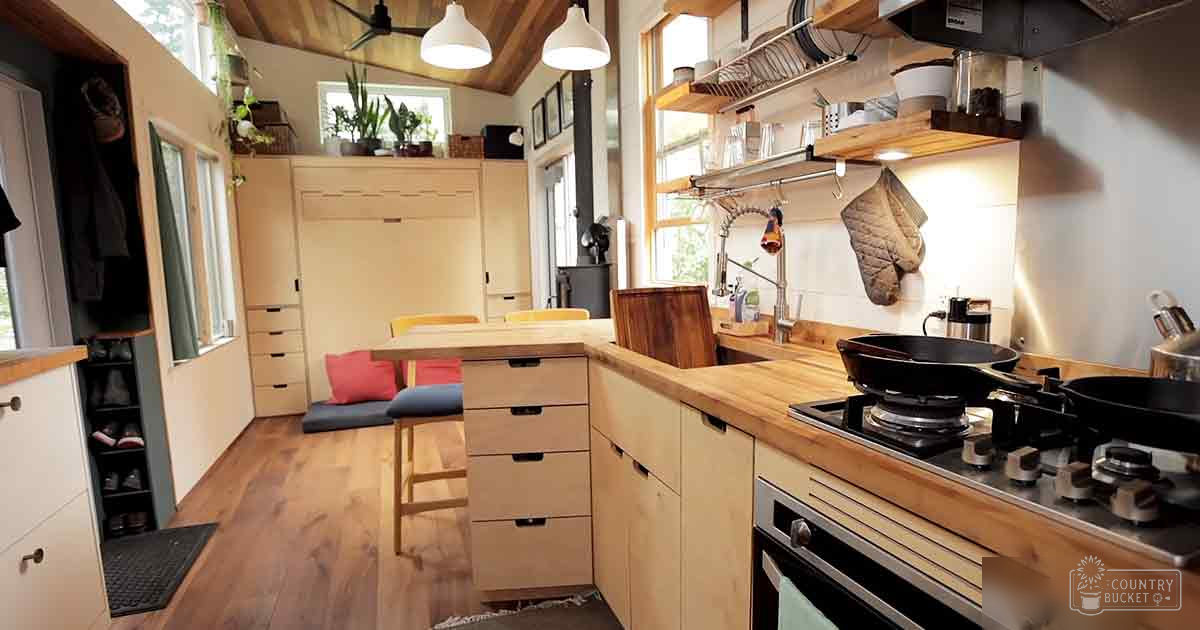 A cozy kitchen area with wooden cabinets, a gas stove, dining table, and natural light from windows, suggesting it's daytime during spring or summer.