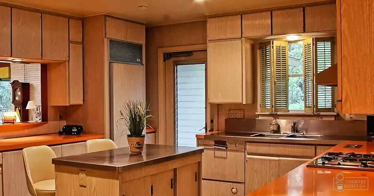 A residential kitchen scene with wooden cabinets, a central island with a dark countertop and potted plant, chairs at the island, a clock on the wall, lamp, radio or speaker unit near refrigerator, small items on countertops, stove, oven or dishwasher under sink; window shutters partially open allowing natural light in, greenery outside suggesting spring or summer
