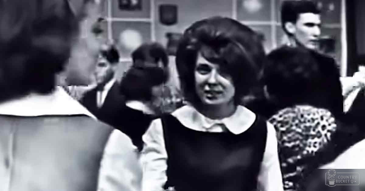 A young adult female in a school setting, wearing a black dress with white collar and light sleeves, surrounded by other students; lockers and bulletin boards suggest an everyday school environment.