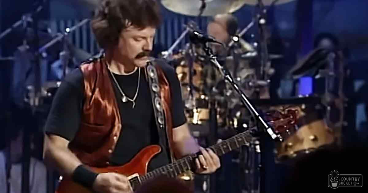 A middle-aged male guitarist in a black short-sleeved shirt with a red vest over it, performing live music on stage at an indoor venue.