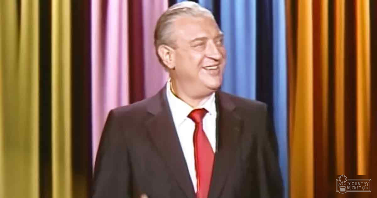 An older adult male in a formal setting with a dark suit jacket, bright red tie, and collared shirt, standing against colorful striped curtains.