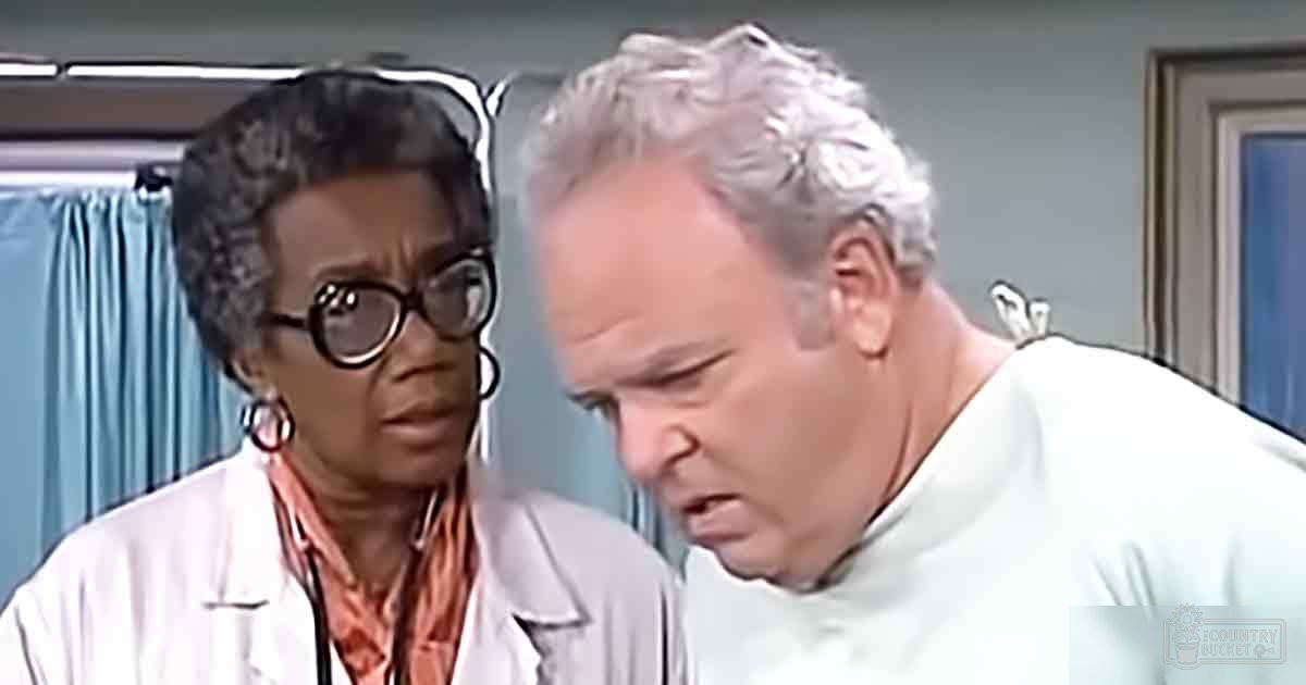 An older adult female healthcare professional wearing a white lab coat and an orange patterned shirt, with large hoop earrings, interacts with another middle-aged to older adult male in a medical setting with light blue curtains and framed artwork.
