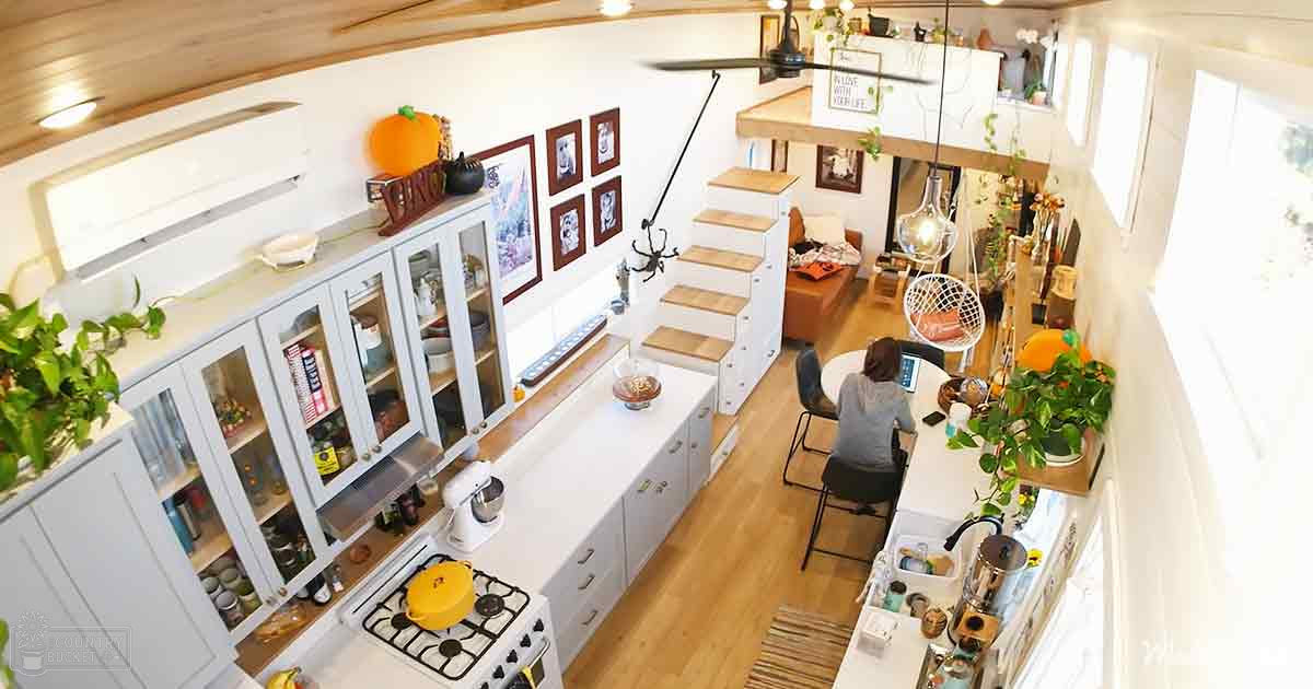 A modern kitchen interior with an adult female seated at a dining table using a laptop; white cabinets, glass doors, gas stove, wooden staircase, framed artwork, hanging plants, ceiling fan, small round chair, various appliances, natural light from large windows or glass doors on the right side of the image