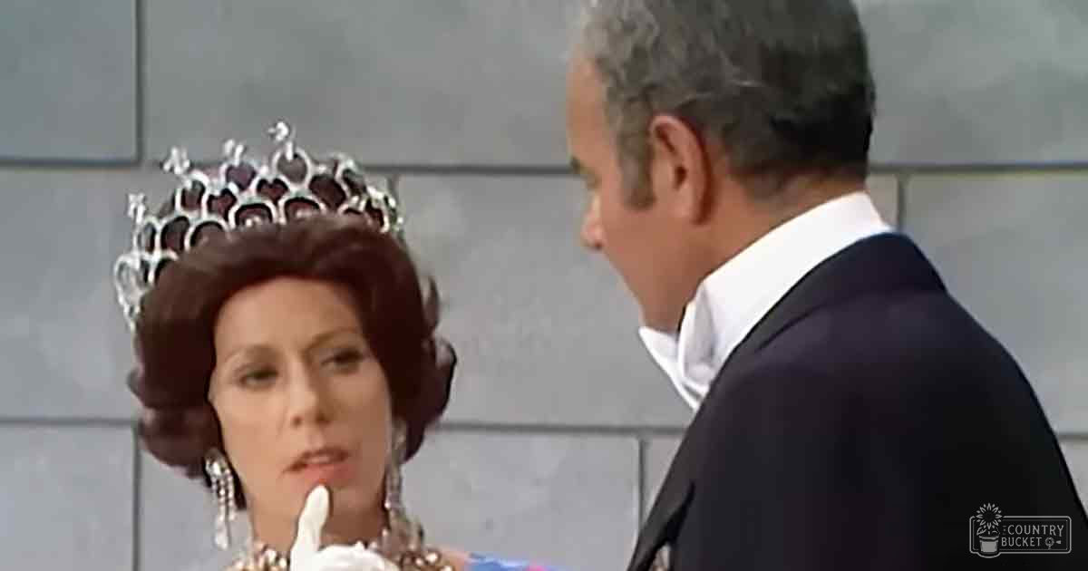 A woman in formal attire with a tiara, wearing an elaborate hairstyle and large earrings, accompanied by a middle-aged to older adult man in a dark suit. The setting appears to be indoors, possibly at a hotel lobby or event venue.