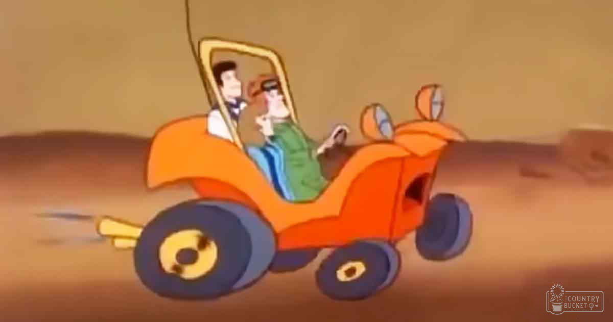 An orange off-road buggy in motion, driven by an adult male with short dark hair wearing a green jacket over a blue shirt or sweater and a white-shirted passenger with short brown hair and a black tie, set against a bright midday desert landscape.