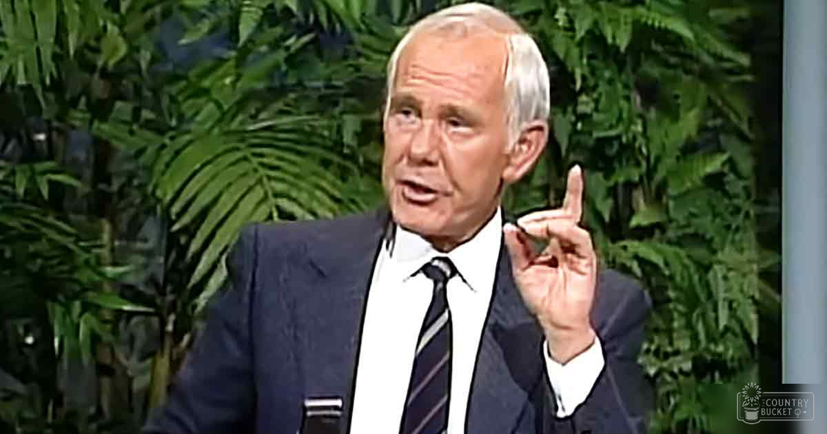 An older adult male in a dark suit, white striped tie, in a studio setting with green foliage backdrop.