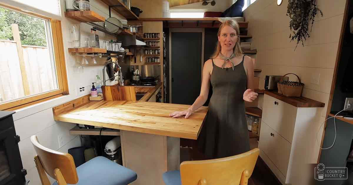 Middle-aged female in a kitchen setting, wearing a sleeveless dark gray knee-length dress with thin straps and tie-like neckline detail, surrounded by wooden furniture and greenery visible through the window.