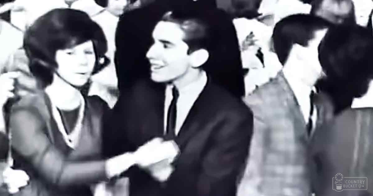 A young adult male in a dark suit jacket at an indoor social gathering with multiple people around him.