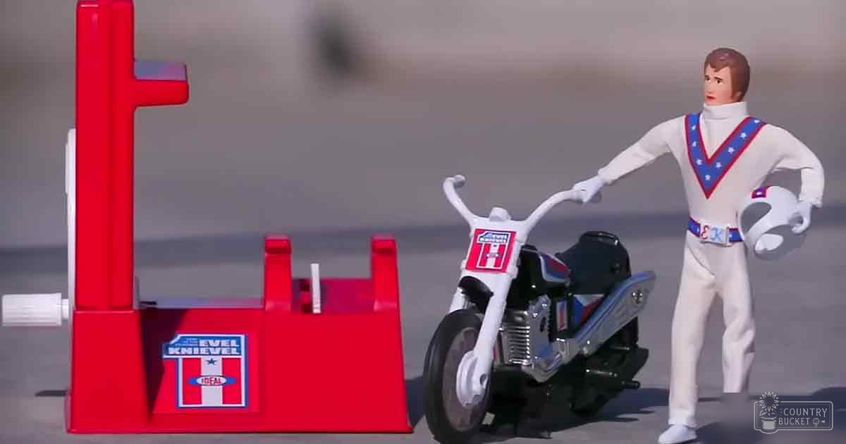 A toy figure resembling Evel Knievel, dressed in a white jumpsuit with blue stars and stripes, is positioned on an 'EVEL KNIEVEL' ramp. The scene includes a red ramp, a white motorcycle labeled 'Ideal,' all displayed indoors for collectible purposes.