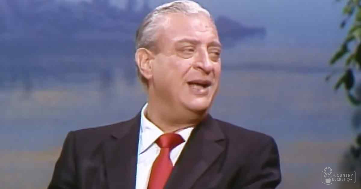 An older adult male in a tailored suit with a red tie, standing outdoors near water under natural daylight.