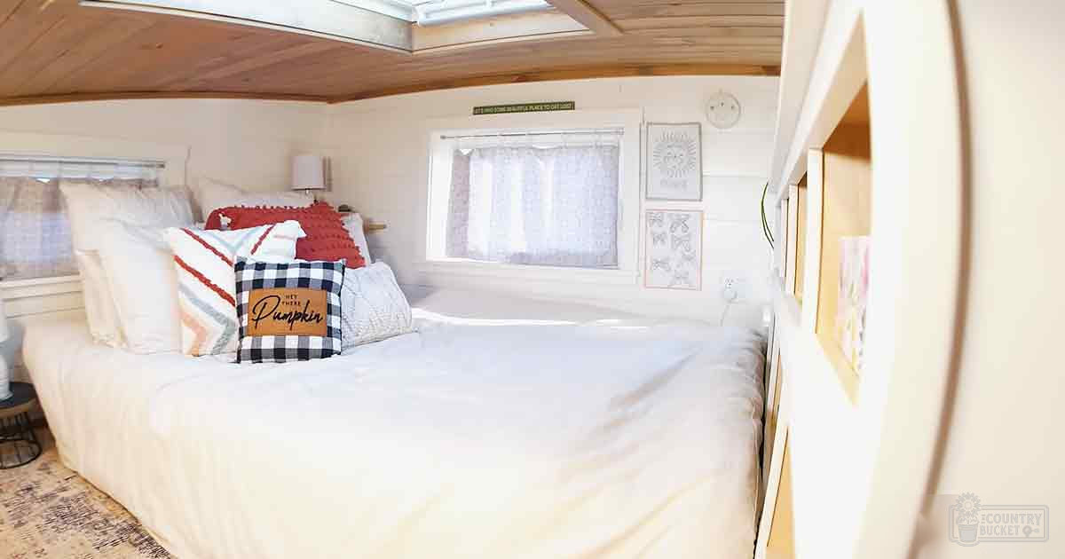 Small bedroom setup with white bedding, wooden ceiling and flooring, natural light from windows, and framed wall art in an RV or small cabin.