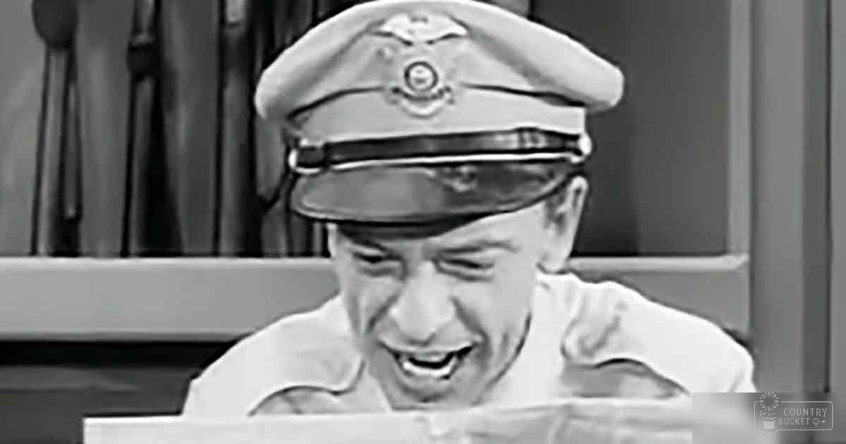 A man in a white military uniform with a peaked cap adorned with insignia stands indoors near a wooden chair, natural daylight illuminates the scene.