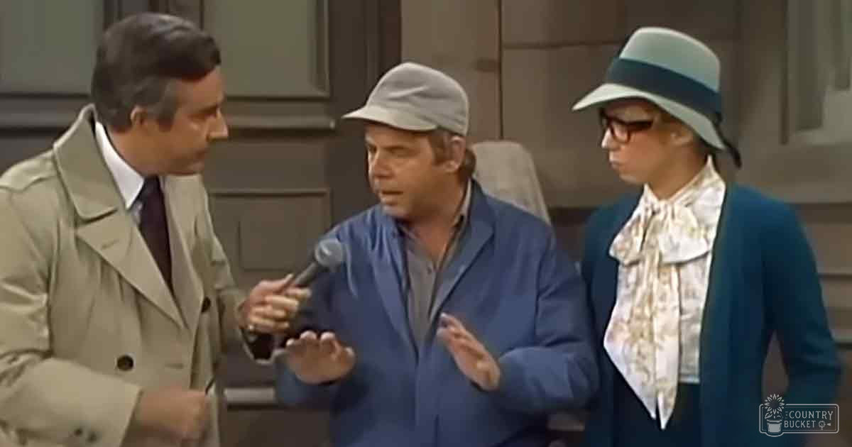 Middle-aged man in a beige trench coat with a white shirt and dark tie stands outdoors near an interview or recording setup involving microphones held by another middle-aged individual wearing a blue jacket over a gray shirt. A woman nearby wears a floral scarf, wide-brimmed hat, and sunglasses.