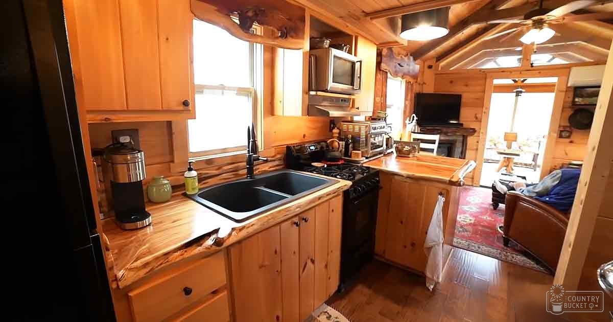 Rustic cabin kitchen with double sink, wooden countertops, coffee maker, stove, microwave, small television, and floral-patterned red rug leading into another room.