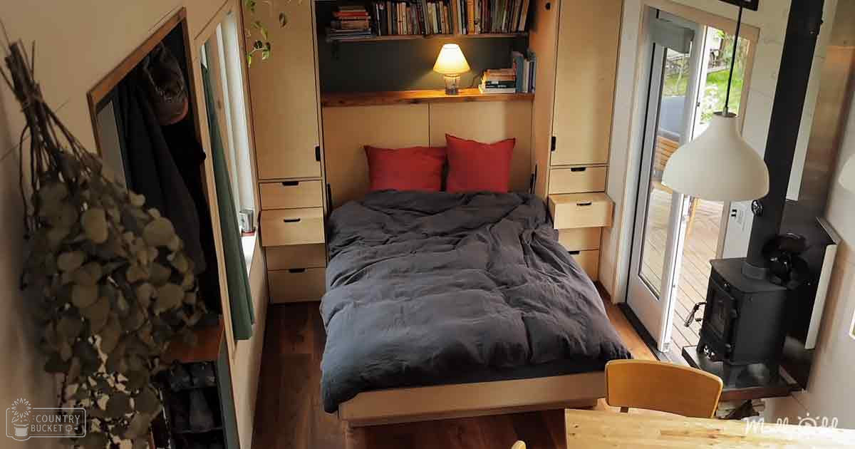 A cozy bedroom scene with dark gray bedding, red pillows, wooden furniture, and a wood-burning stove in a small house or cabin, bathed in natural daylight.
