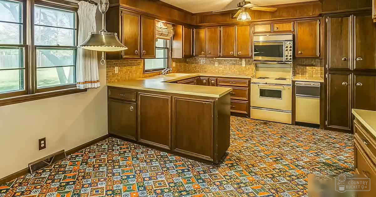 A residential kitchen with wooden cabinets, a central island, patterned linoleum floor, and natural light from windows. The setting appears to be indoors within a home.
