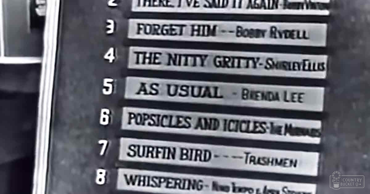 A mid-20th century music chart displayed indoors, with song titles and artist names against a black-and-white background.