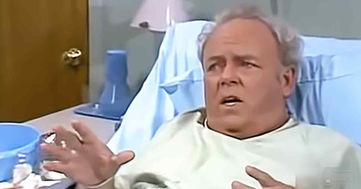A middle-aged adult male wearing a white hospital gown stands in an indoor hospital room, illuminated by natural light.