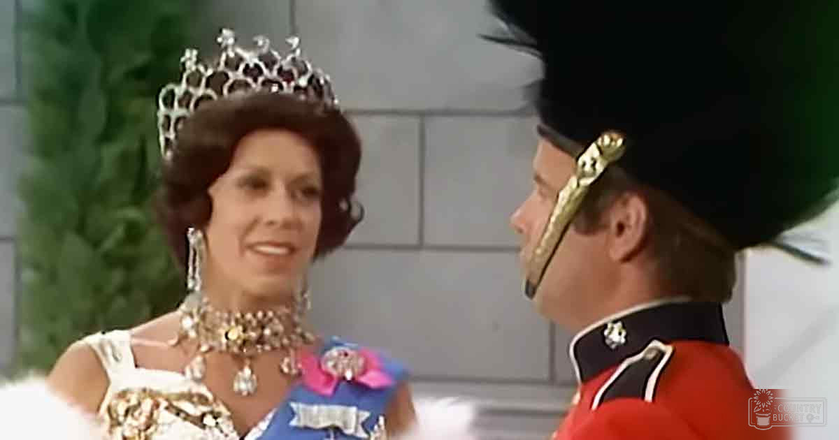 A woman in a formal setting with a white dress and blue sash, wearing a large tiara and long earrings. A man in a red military uniform stands beside her.
