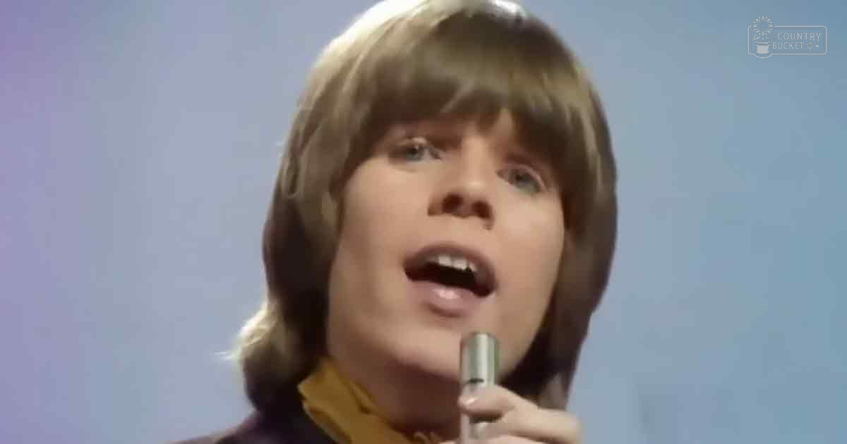 A young adult male singer wearing a mustard yellow 1970s-era garment with a high collar, singing into a vintage-style microphone in an indoor performance setting.