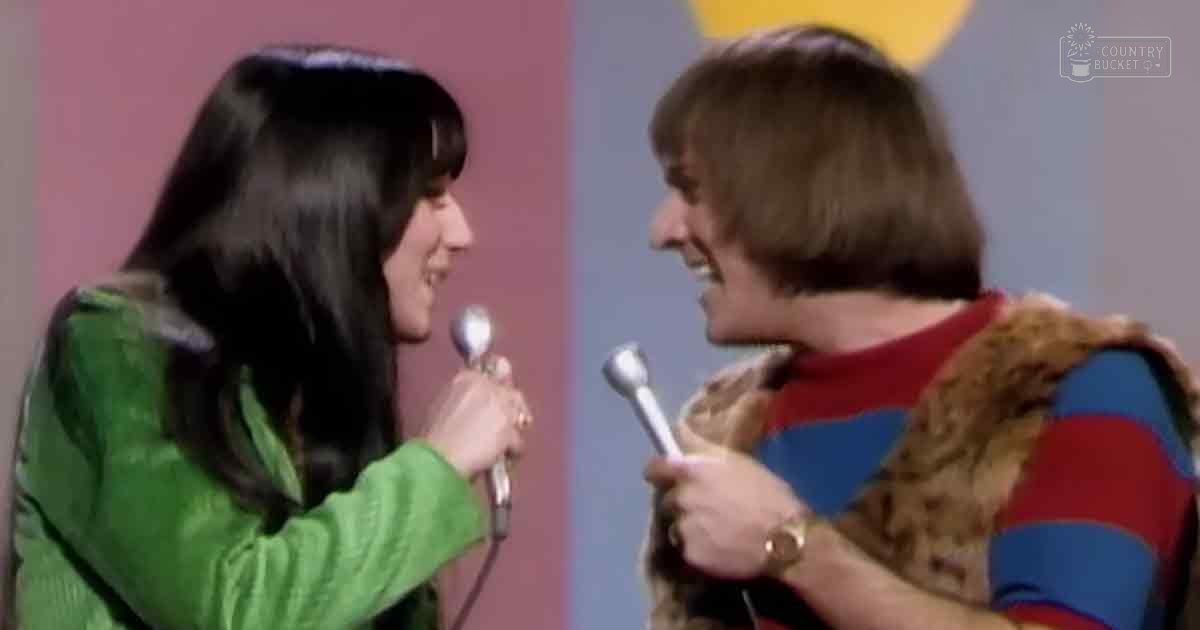 A young adult female in a green, long-sleeved, high-necked garment and a middle-aged male in a red and blue striped shirt with a fur-like vest perform on stage against a backdrop of pink and blue panels under studio lighting.