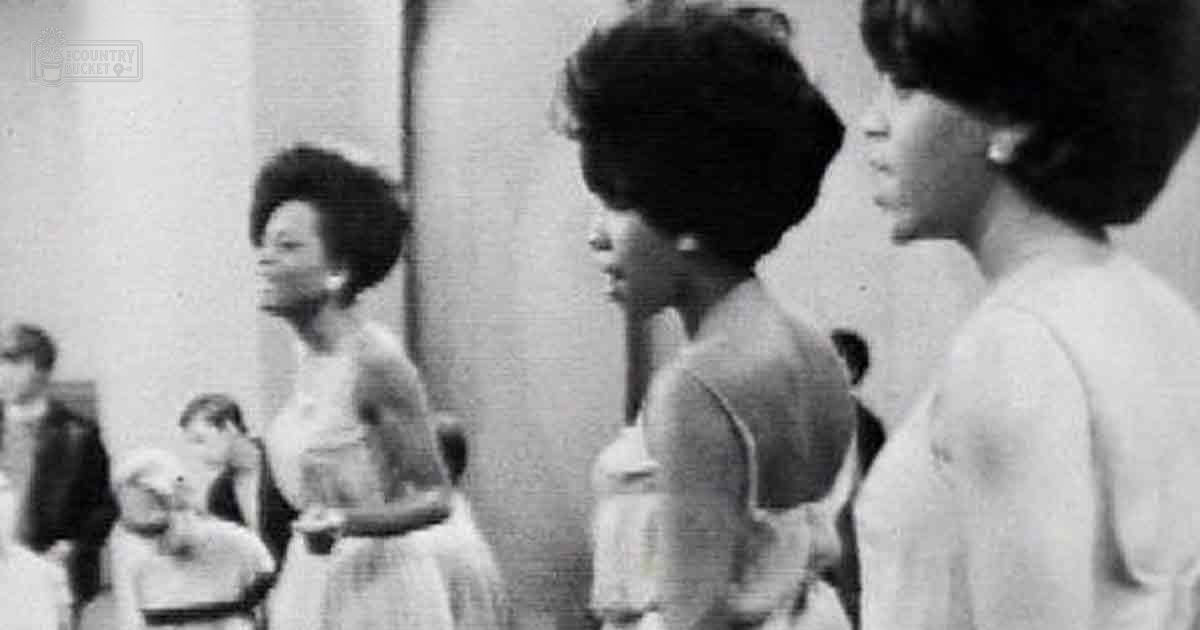 Two adult females in a mid-20th-century indoor setting with light-colored garments; one facing away wearing a sleeveless dress, another partially obscured with an afro-style hairdo. A child dressed in white is also present.
