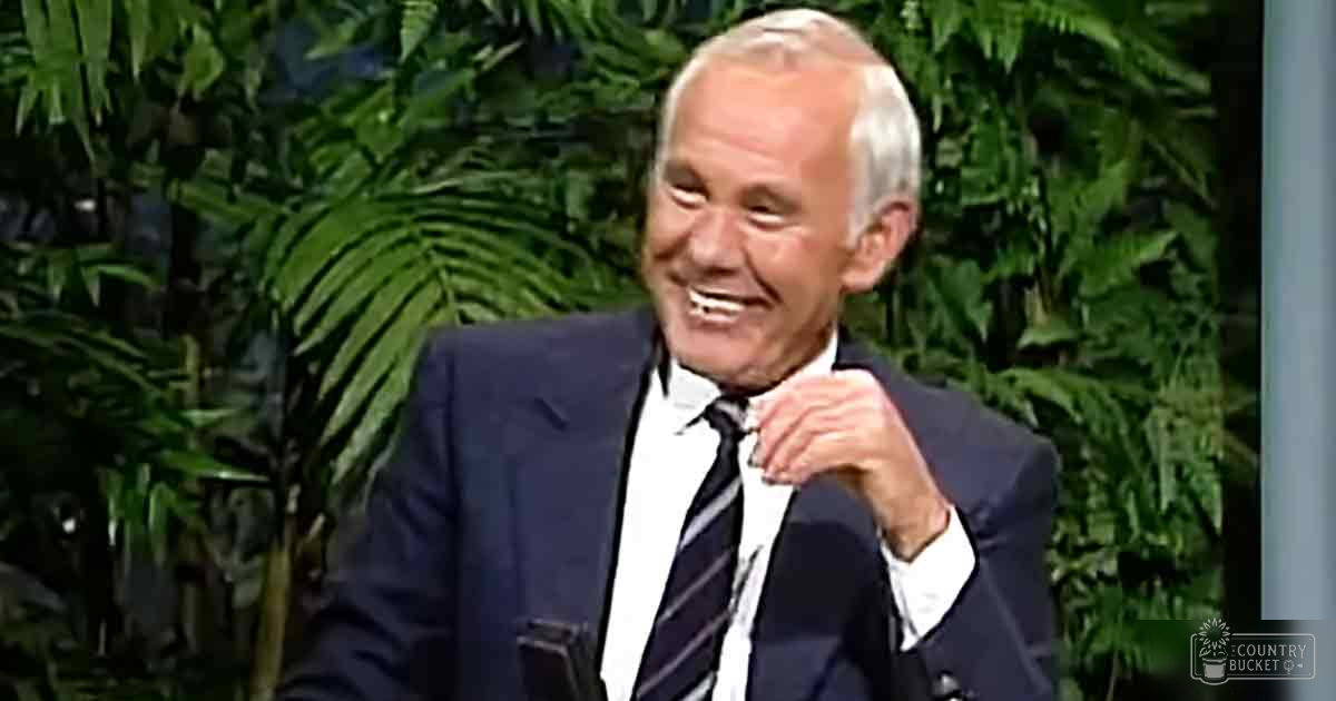 An older adult male wearing a dark blue suit jacket, white dress shirt, and striped tie in an indoor setting with lush green foliage and studio lighting.