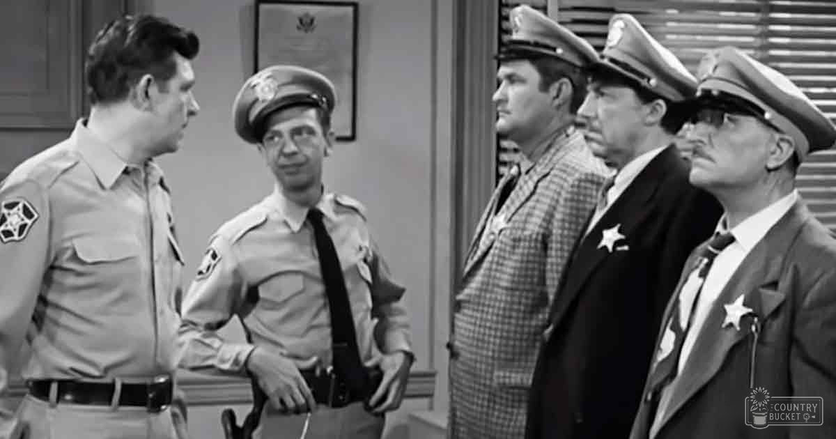 A group of law enforcement officers in an indoor setting, possibly a police station; uniform shirts with epaulets and badges, plaid shirt, dark suit jacket; framed certificate on the wall.
