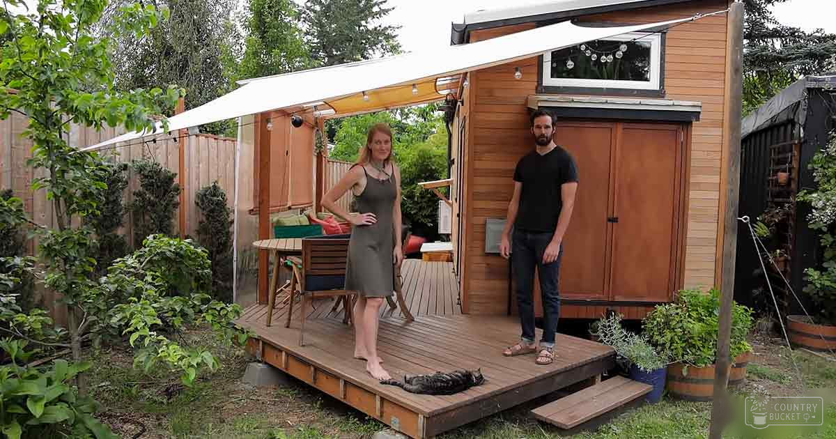 A couple stands in front of their tiny house in a backyard garden setting during spring or summer with soft afternoon light.